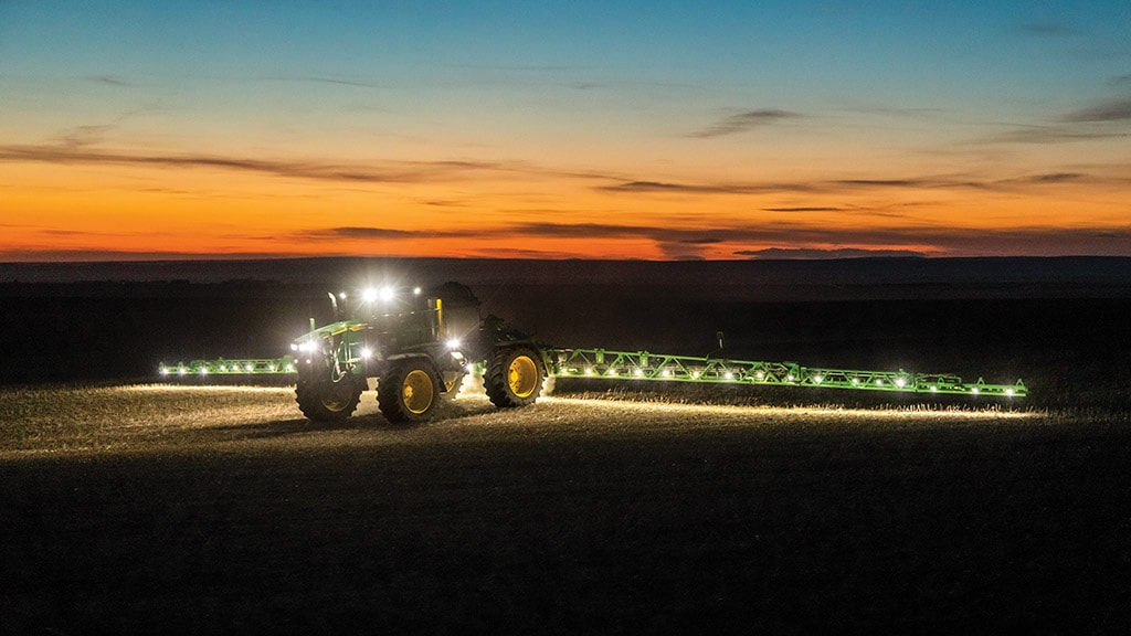 A John Deere Self-Propelled Sprayer with lighted booms extended while applying in a dark, fallow field at nighttime.